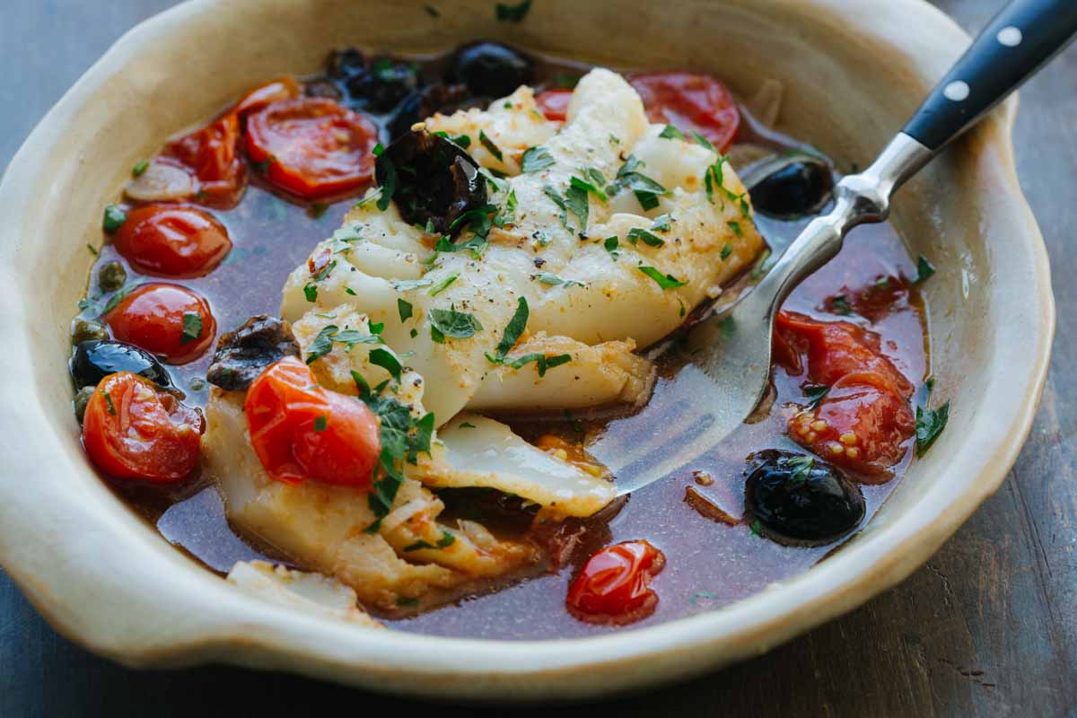 Fork lifting a piece of flaky white fish from a bowl of acqua pazza, showing the tomato-olive broth and fresh herbs.