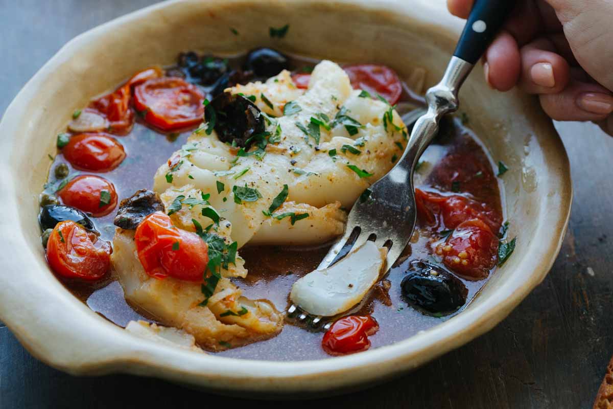 Hand holding a fork taking a bite of fish acqua pazza, with tomatoes and olives visible in the broth below.