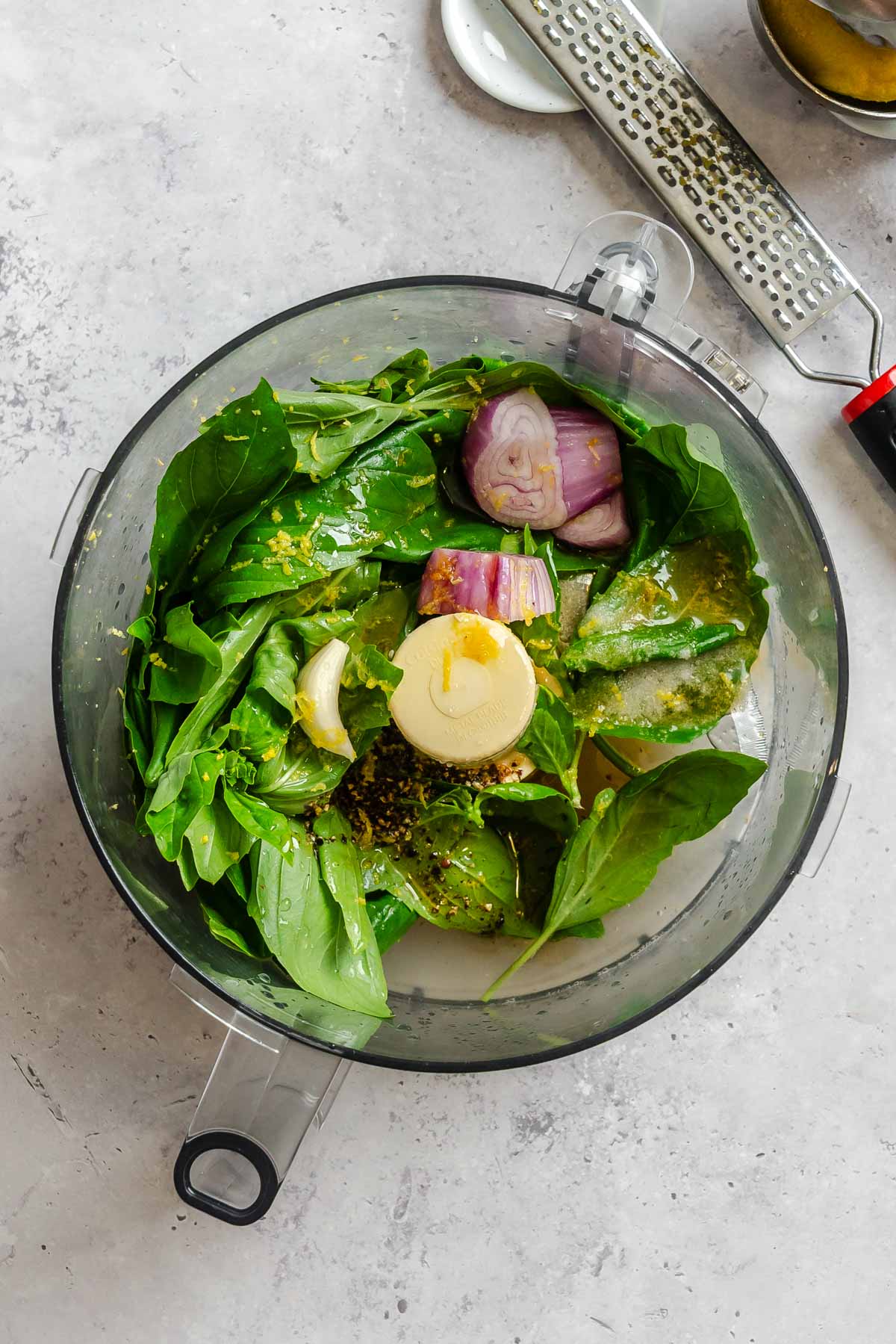Basil leaves, lemon zest, shallot, and garlic in the bowl of a food processor before blending.