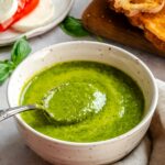 Close-up of basil vinaigrette in a bowl with a spoon resting inside, grilled bread and a caprese plate in the background.