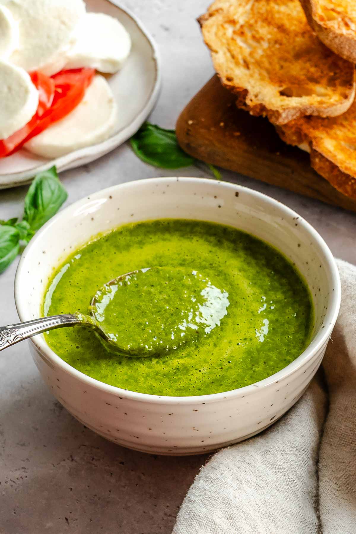 Close-up of basil vinaigrette in a bowl with a spoon resting inside, grilled bread and a caprese plate in the background.