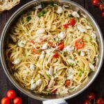 Overhead shot of linguine with crab and cherry tomatoes in a stainless steel skillet, surrounded by crusty bread, fresh tomatoes on the vine, and shallots.