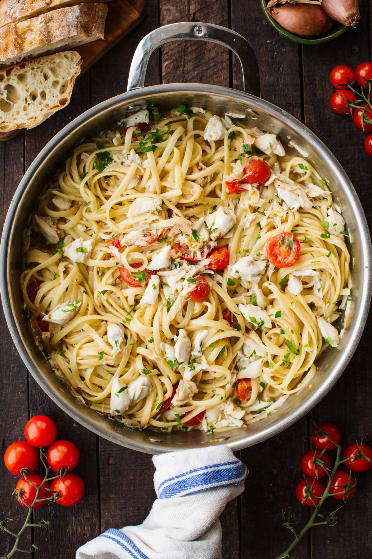 Overhead shot of linguine with crab and cherry tomatoes in a stainless steel skillet, surrounded by crusty bread, fresh tomatoes on the vine, and shallots.