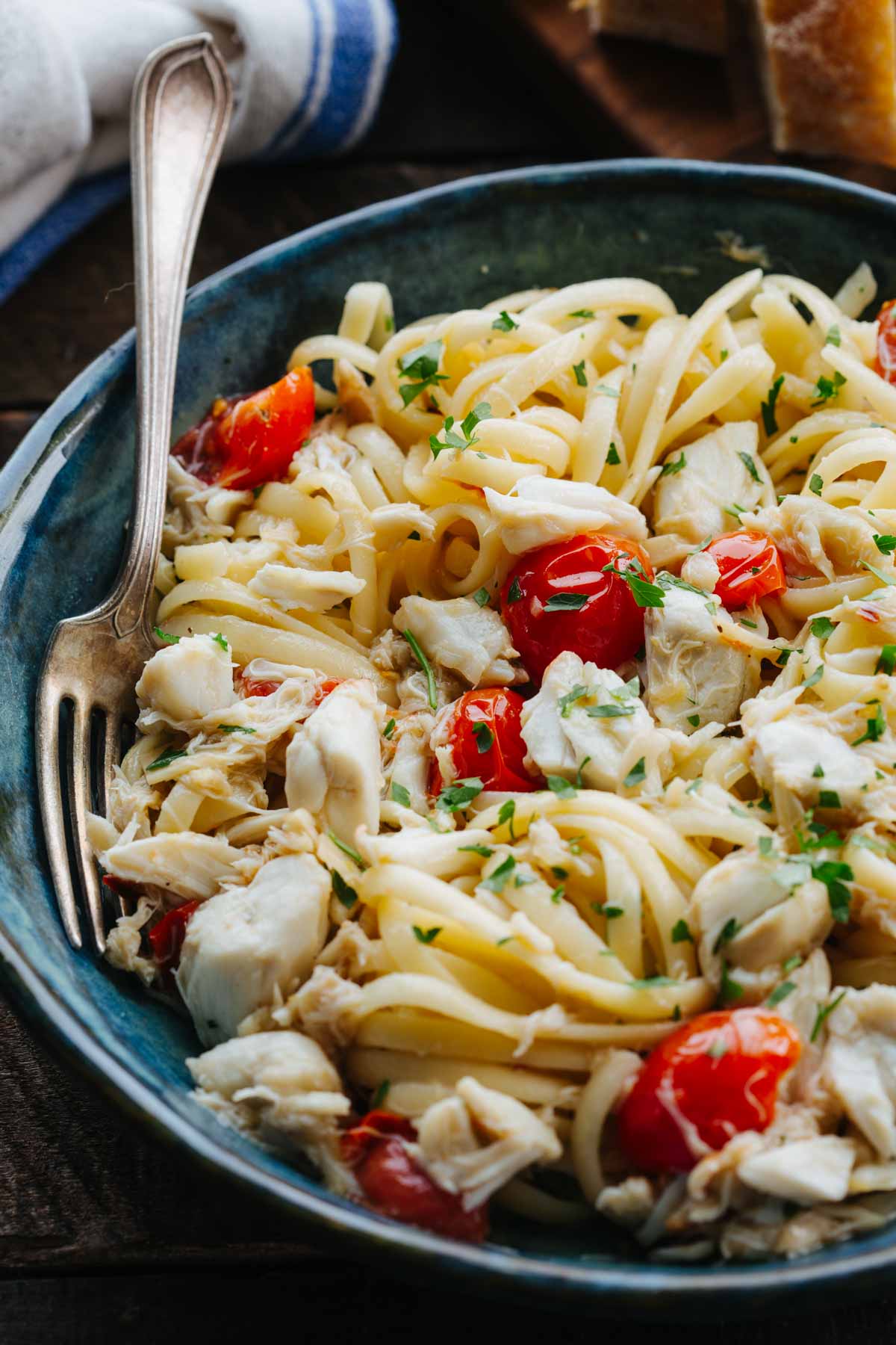 A blue ceramic bowl of crab linguine with cherry tomatoes and parsley, fork resting on the edge."