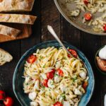 Overhead view of a bowl of crab pasta beside the skillet it was cooked in, with sliced ciabatta, garlic, and tomatoes on the side.