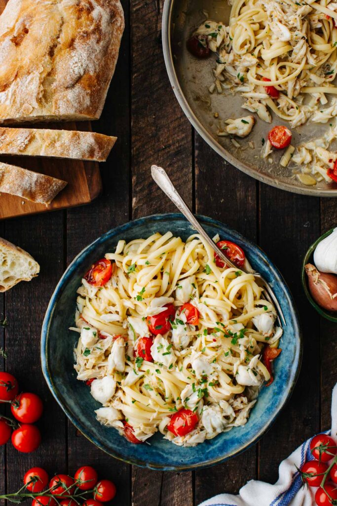 Overhead view of a bowl of crab pasta beside the skillet it was cooked in, with sliced ciabatta, garlic, and tomatoes on the side.