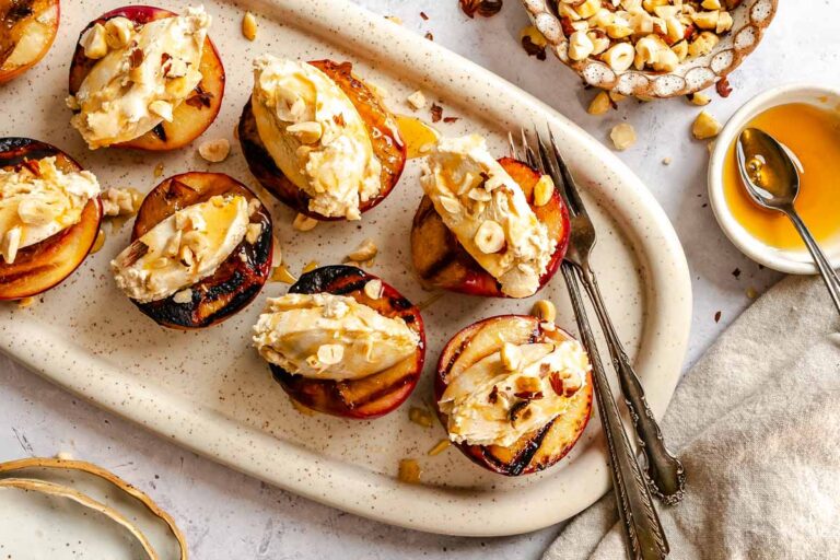 Overhead shot of grilled peach halves on an oval platter, each topped with mascarpone cream, chopped hazelnuts, and a drizzle of honey, with forks resting alongside.