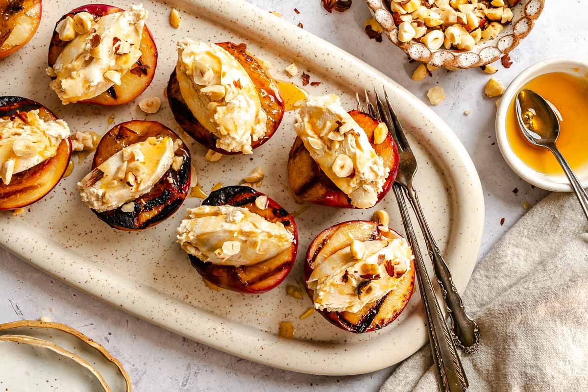 Overhead shot of grilled peach halves on an oval platter, each topped with mascarpone cream, chopped hazelnuts, and a drizzle of honey, with forks resting alongside.