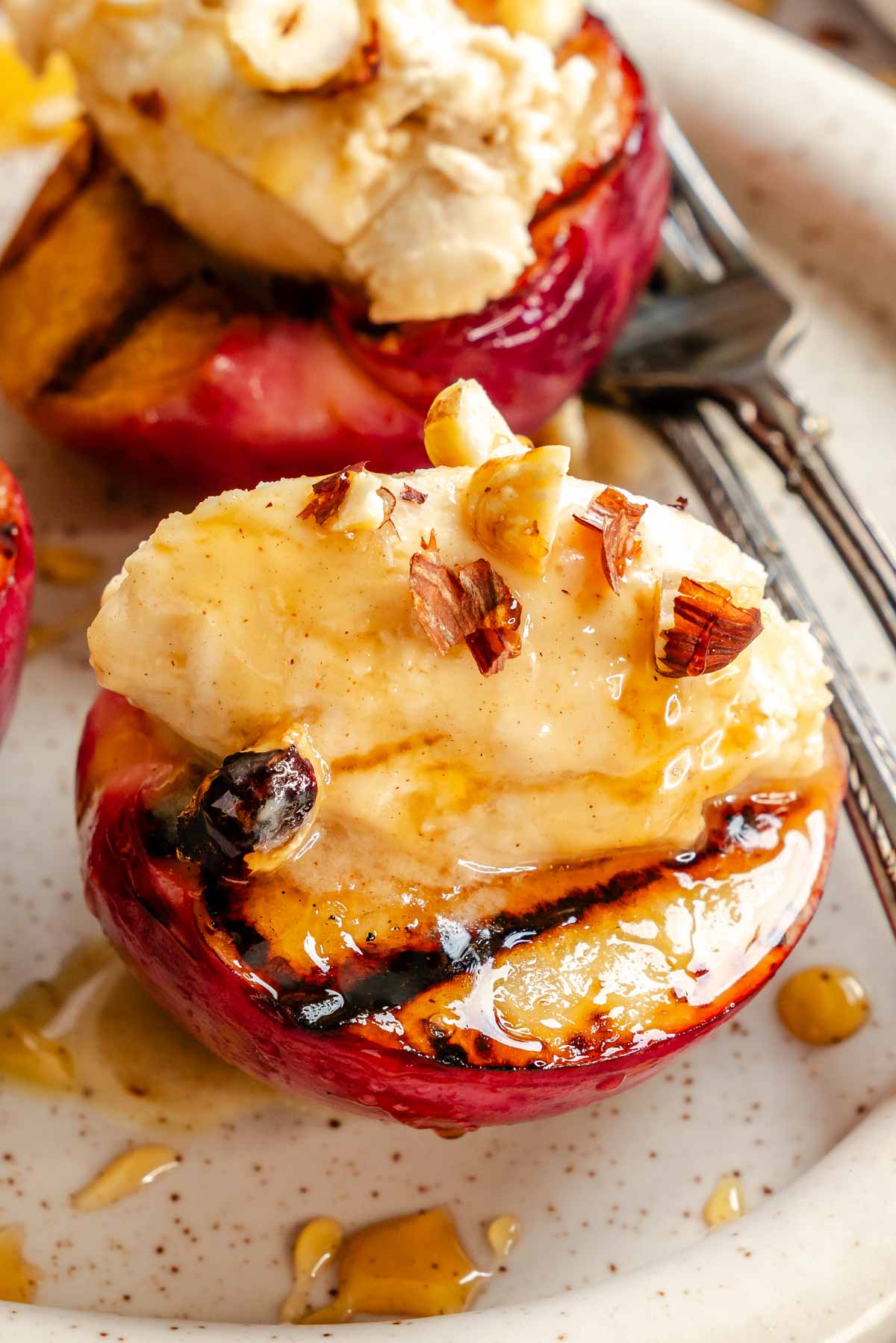 Macro shot of a mascarpone-topped grilled peach half with hazelnut pieces and honey drizzle, showing the charred grill marks on the peach.