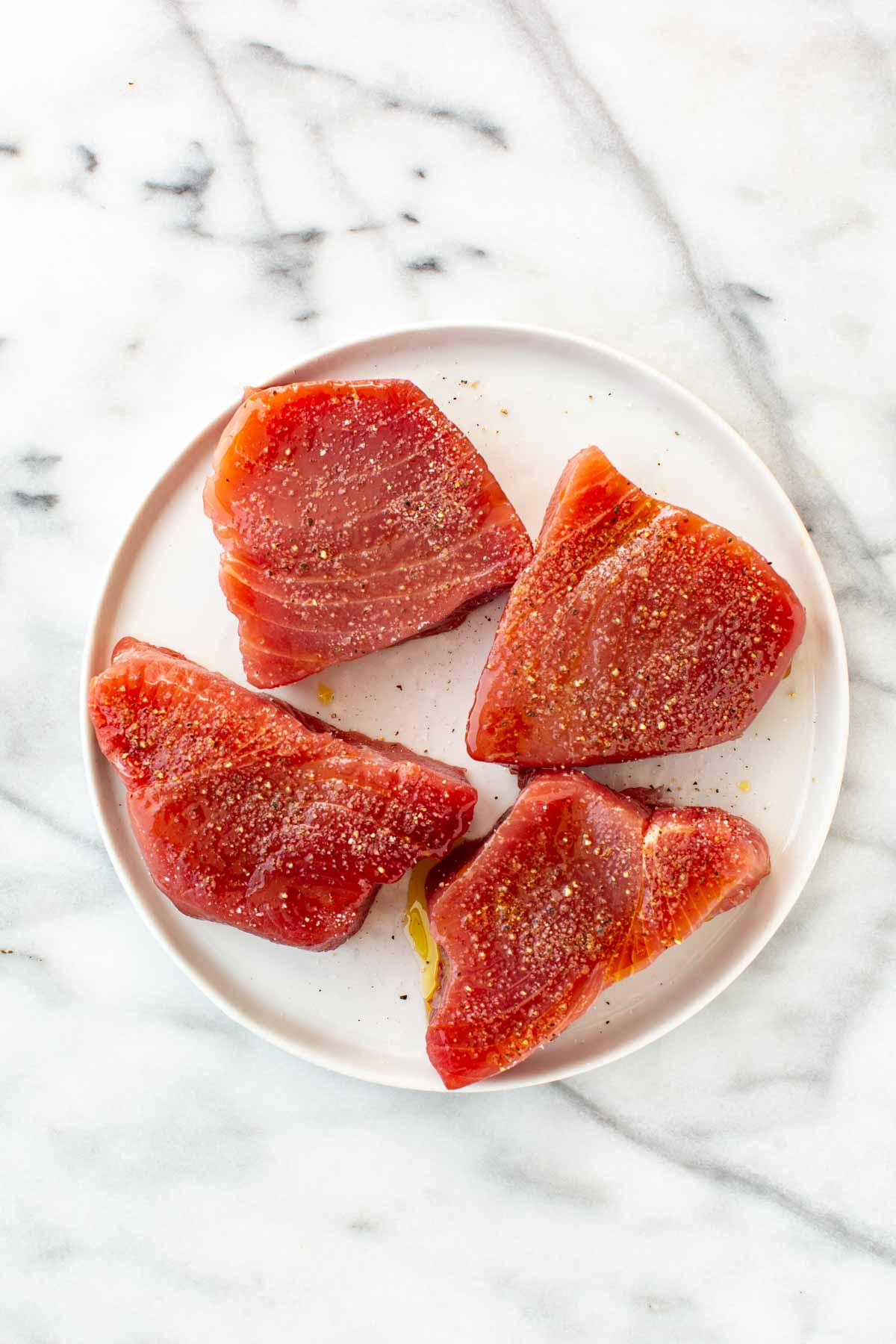 Four raw tuna steaks seasoned with salt and pepper, arranged on a white plate.