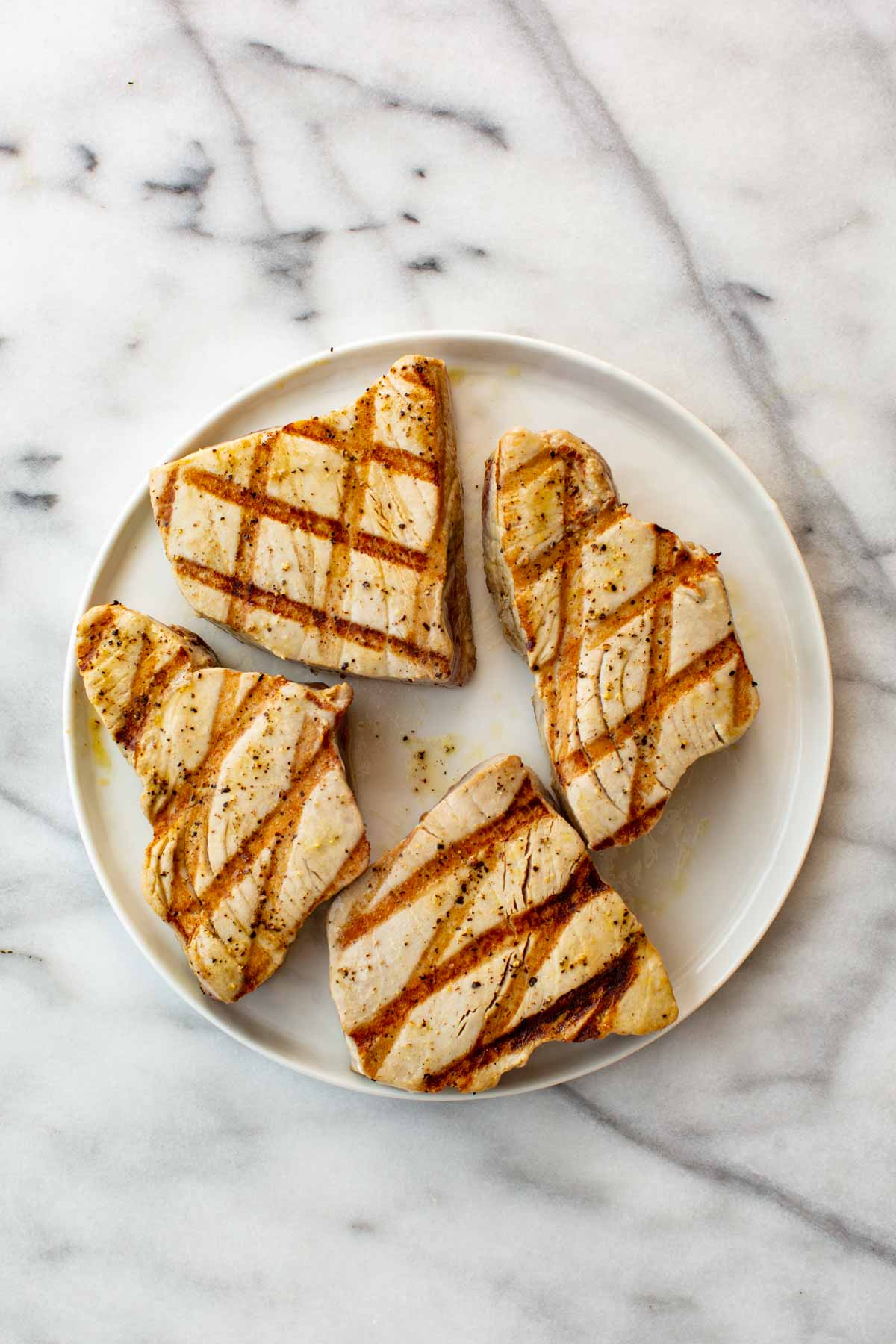 Four tuna steaks with dark grill marks, cooked and resting on a white plate.