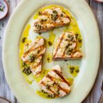 Overhead view of grilled tuna steaks on an oval platter, drizzled with caper vinaigrette and surrounded by charred lemon halves and fresh herbs.