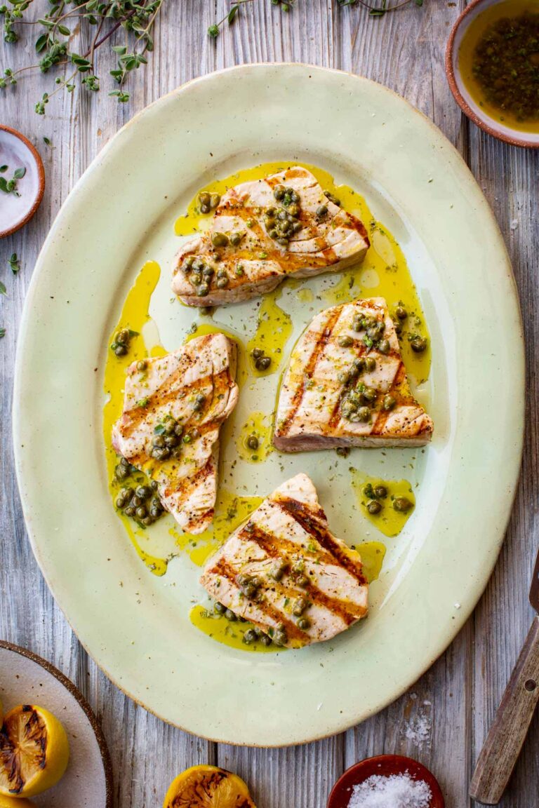 Overhead view of grilled tuna steaks on an oval platter, drizzled with caper vinaigrette and surrounded by charred lemon halves and fresh herbs.