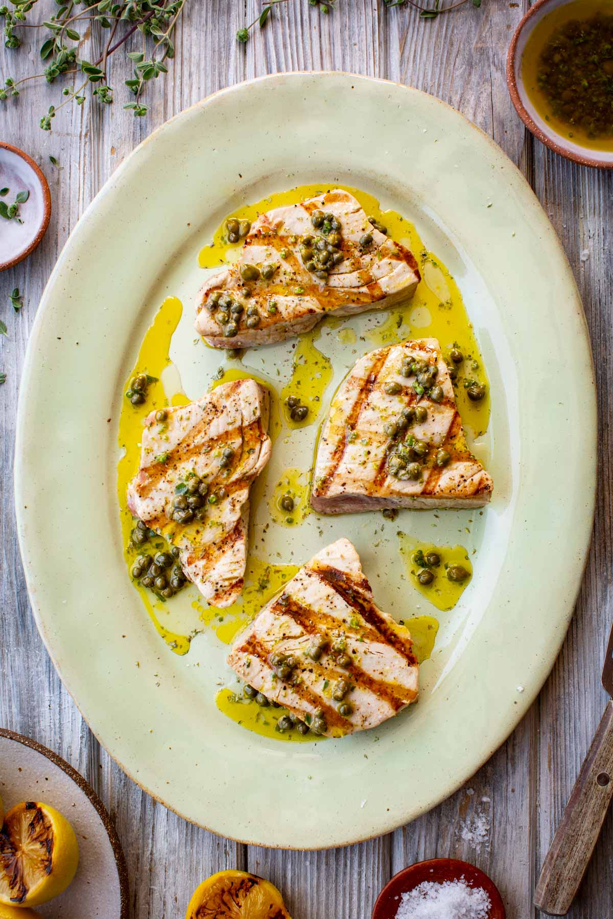 Overhead view of grilled tuna steaks on an oval platter, drizzled with caper vinaigrette and surrounded by charred lemon halves and fresh herbs.
