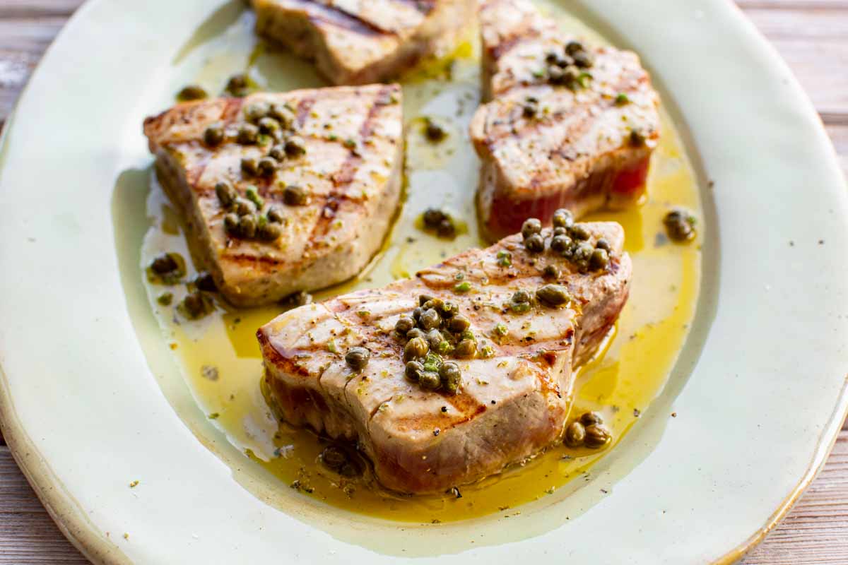 Close-up of grilled tuna steaks topped with whole capers and olive oil on a light green plate.