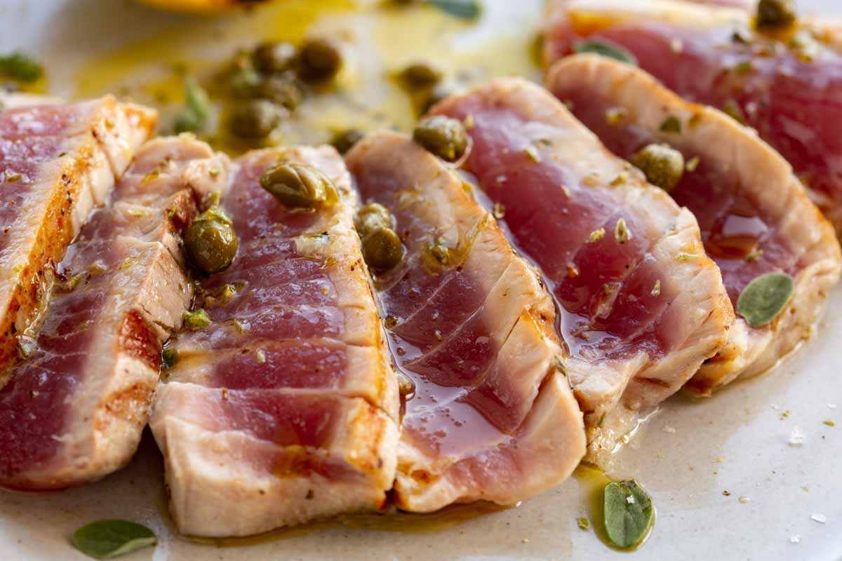 Close-up of sliced grilled tuna showing a pink, rare center, topped with capers and herb oil.