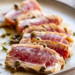 Side angle close-up of sliced grilled tuna steaks with a rosy interior, garnished with capers and glistening with vinaigrette.