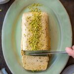 Overhead shot of pistachio semifreddo on a sage green oval platter, being sliced with a knife, topped with crushed pistachios and one cut slice pulled forward.