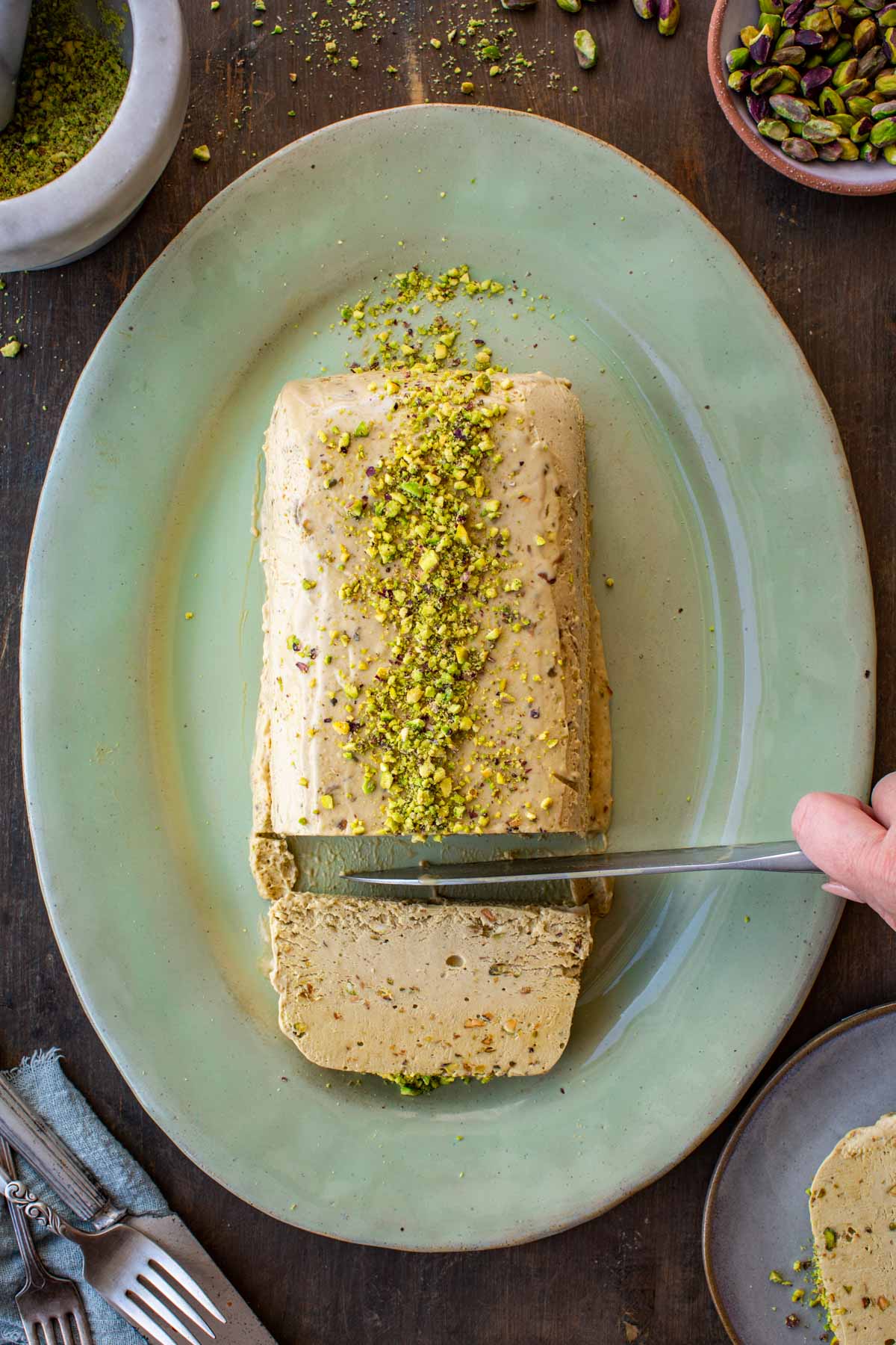Overhead shot of pistachio semifreddo on a sage green oval platter, being sliced with a knife, topped with crushed pistachios and one cut slice pulled forward.