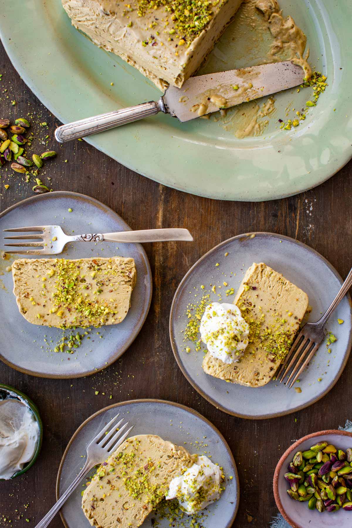 Overhead shot of sliced pistachio semifreddo plated on three individual plates, with the remaining loaf and serving knife on the platter above; two slices topped with whipped cream and crushed pistachios.