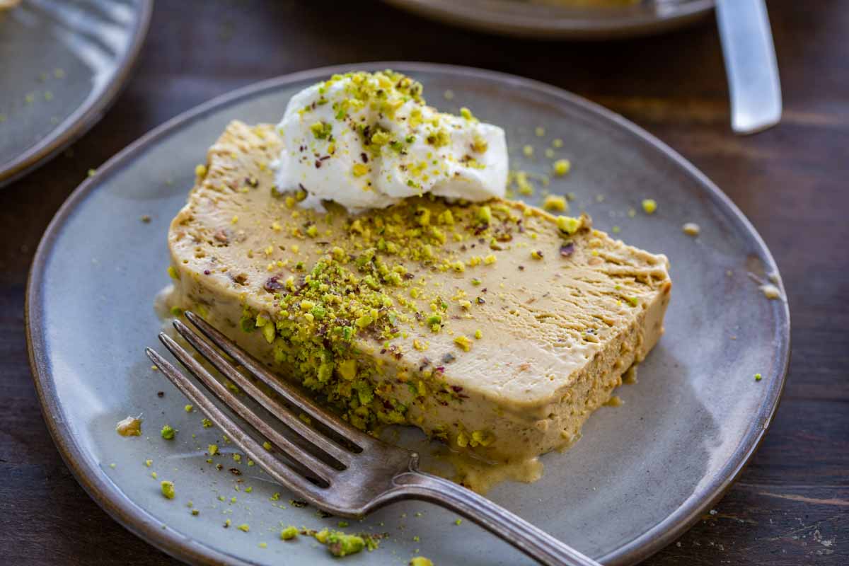 Close-up of a single slice of pistachio semifreddo on a stoneware plate, topped with a dollop of whipped cream and crushed pistachios, fork resting alongside.