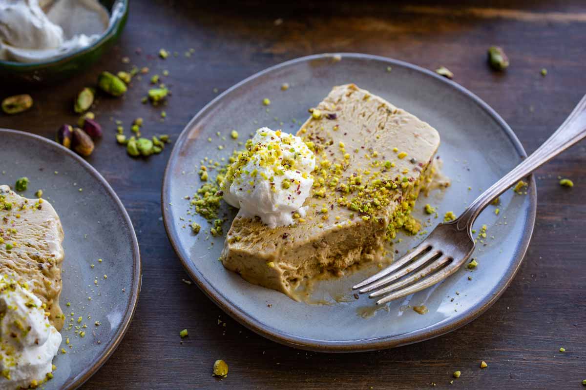 Side angle of a plated pistachio semifreddo slice with whipped cream and crushed pistachios, a second slice visible on a plate in the background.