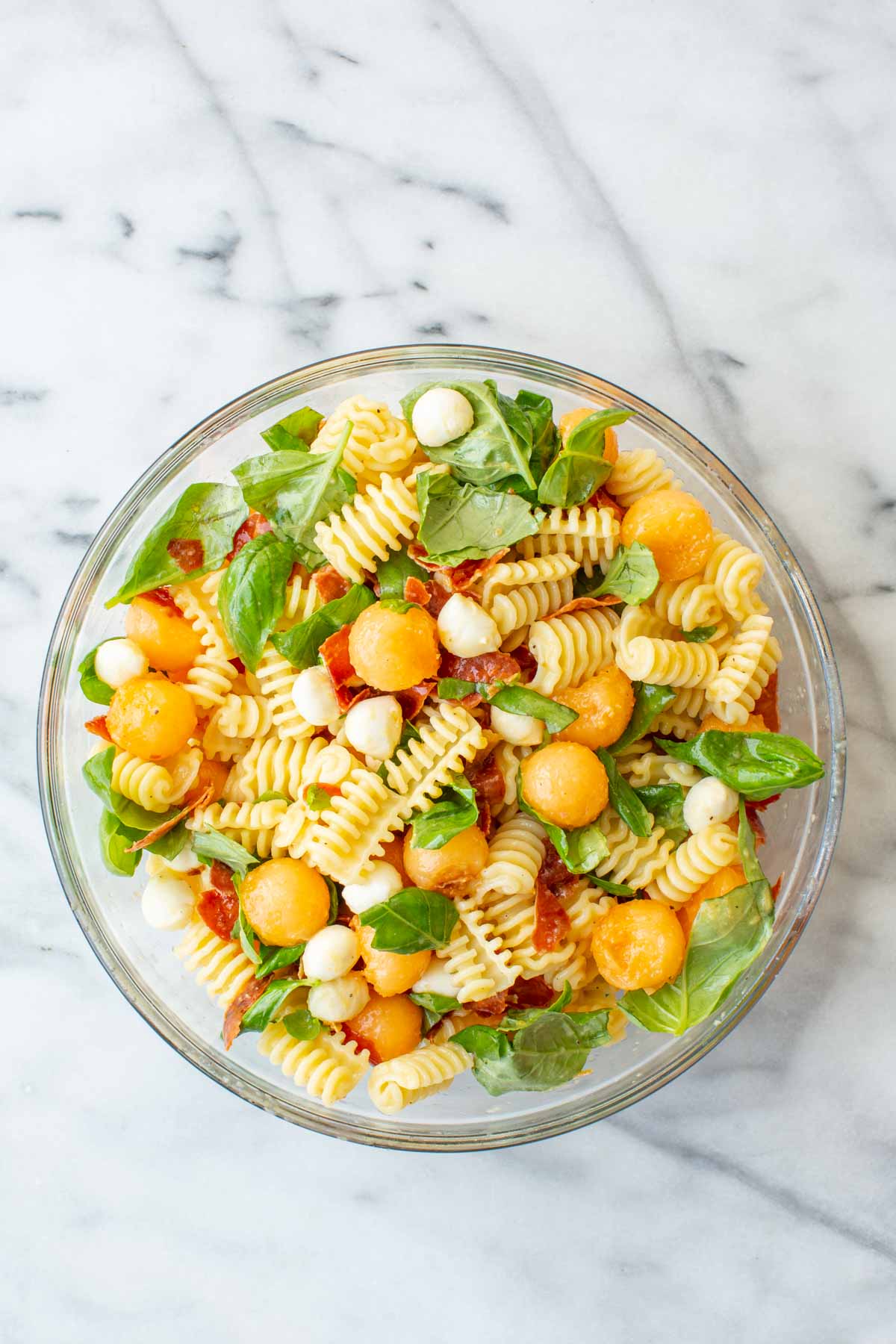 Overhead view of a glass bowl filled with radiatori pasta, cantaloupe balls, mini mozzarella balls, crispy prosciutto pieces, and fresh basil leaves on a marble surface.