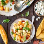 A plated serving of prosciutto melon pasta salad on a speckled ceramic plate with a fork, next to a bowl of mini mozzarella balls, crispy prosciutto shards, cantaloupe wedges, and a yellow linen napkin on a dark wood table.