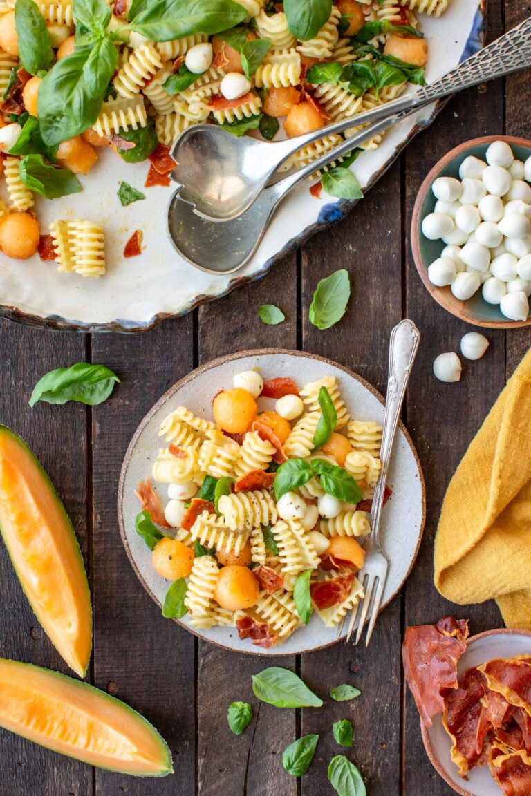 A plated serving of prosciutto melon pasta salad on a speckled ceramic plate with a fork, next to a bowl of mini mozzarella balls, crispy prosciutto shards, cantaloupe wedges, and a yellow linen napkin on a dark wood table.