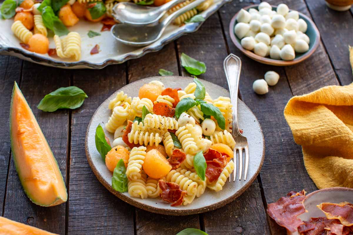 Close-up angled shot of a single serving of pasta salad with melon balls, mozzarella pearls, basil, and crispy prosciutto on a rustic plate with a fork, set on a dark wood table.