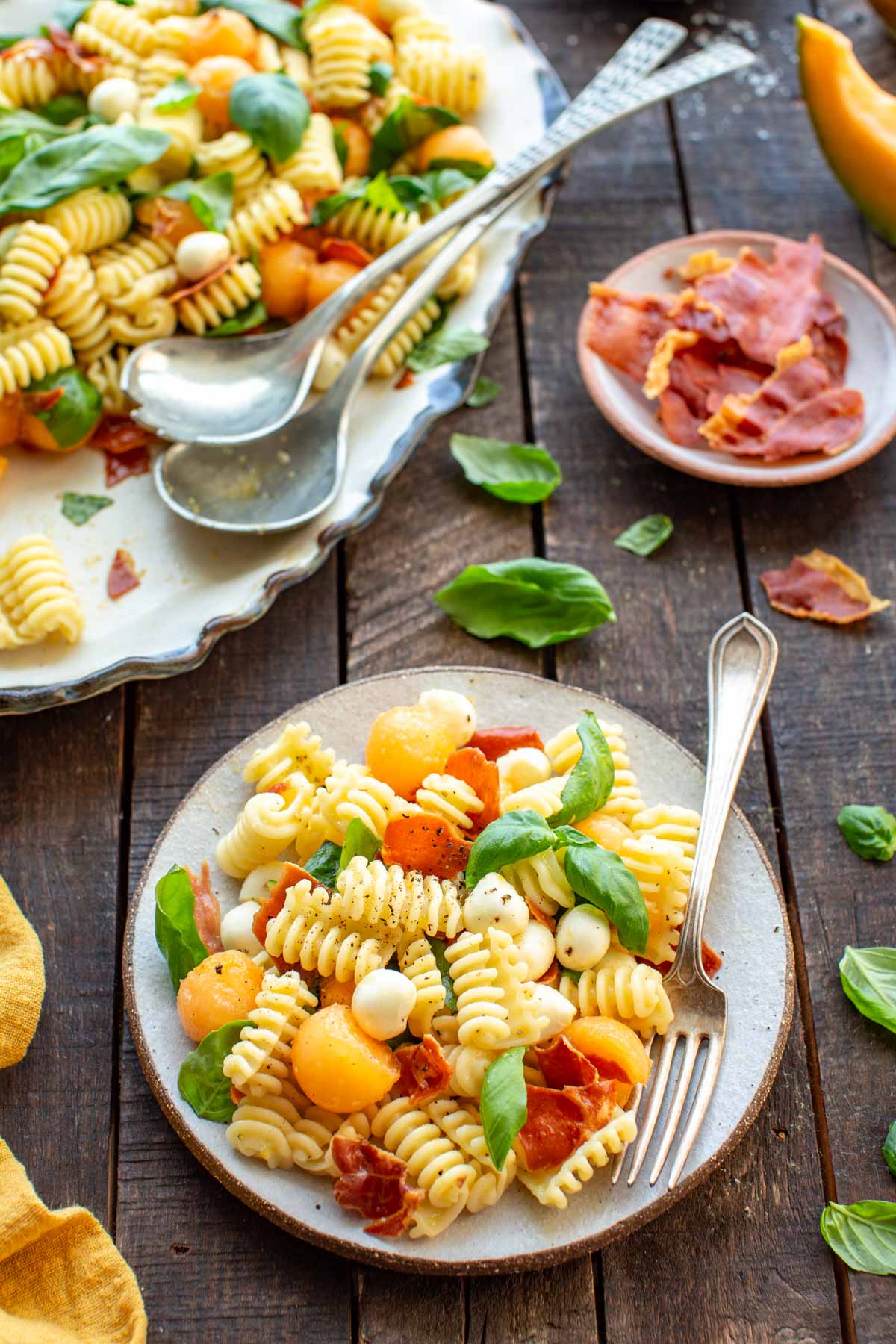 A plated portion of pasta salad with cantaloupe balls, mozzarella pearls, crispy prosciutto, and basil leaves, with a small dish of extra prosciutto pieces and a serving platter in the background.