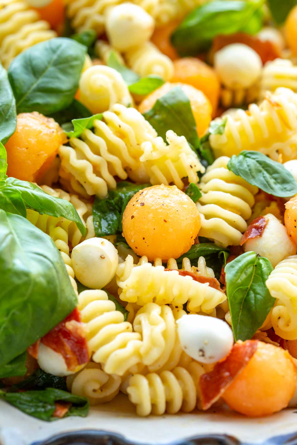 Extreme close-up of radiatori pasta tossed with cantaloupe balls, mini mozzarella, crispy prosciutto, and basil, showing texture and glossy dressing.