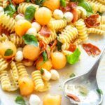 Close-up of pasta salad on a serving platter with a silver serving spoon resting on the edge, showing melon balls, mozzarella, crispy prosciutto, and basil.