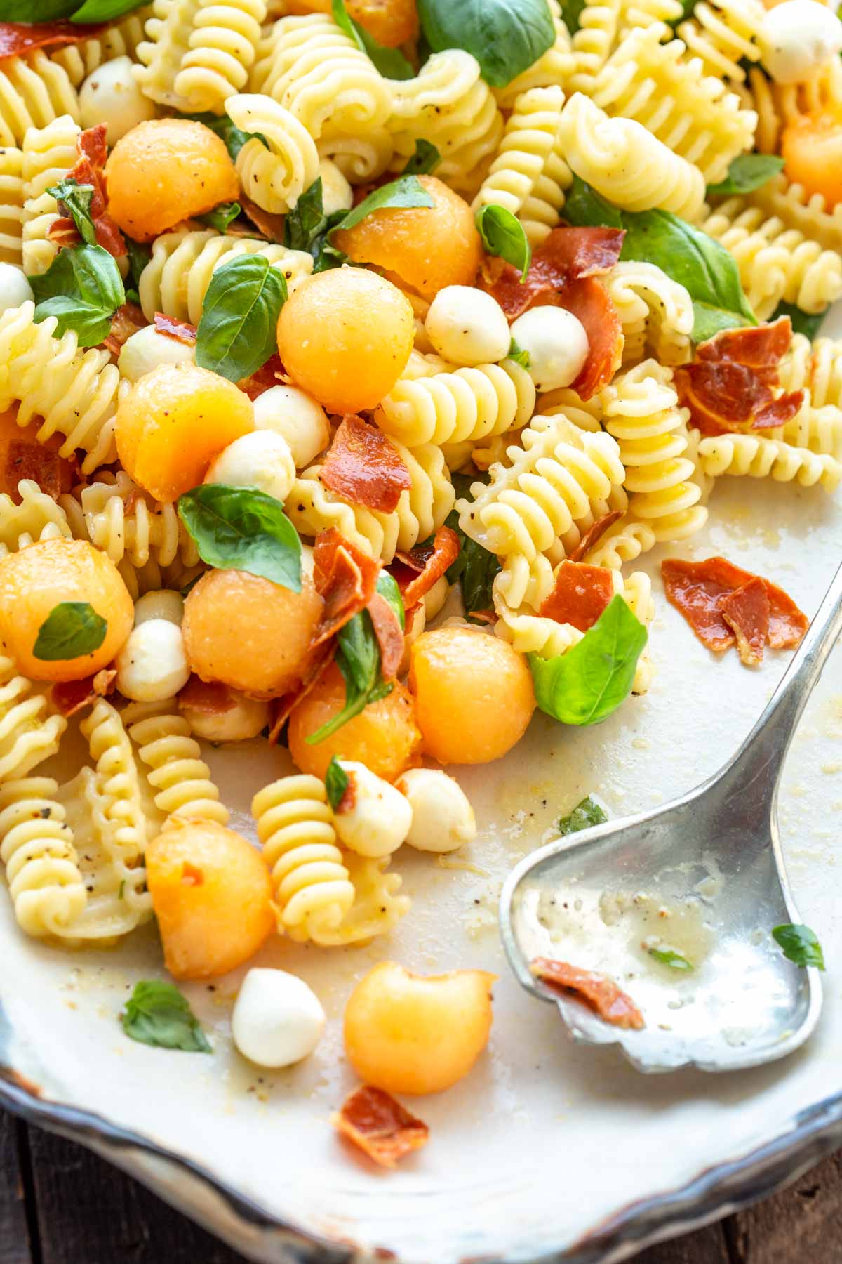 Close-up of pasta salad on a serving platter with a silver serving spoon resting on the edge, showing melon balls, mozzarella, crispy prosciutto, and basil.
