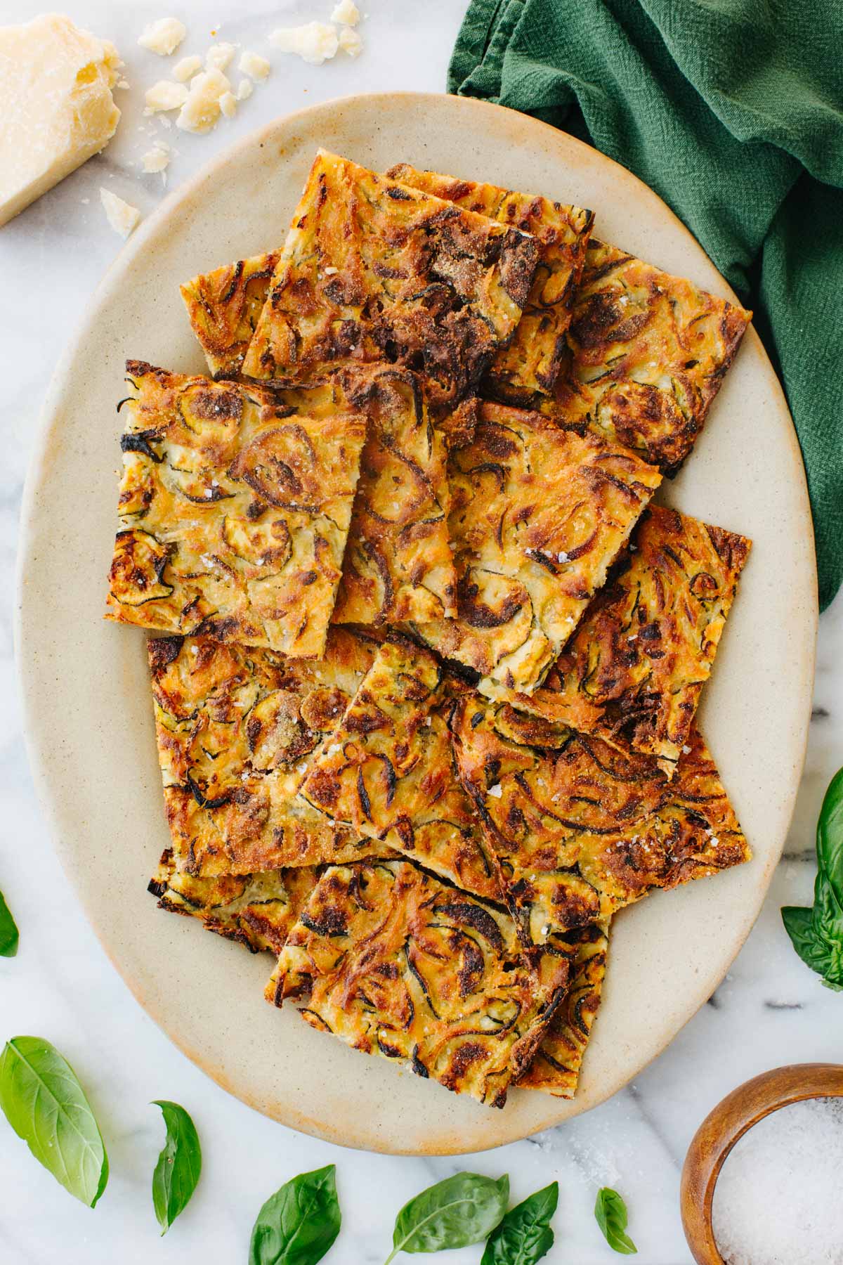 Overhead shot of golden brown zucchini scarpaccia cut into squares and piled on an oval ceramic plate, with shaved parmesan, fresh basil, and a green linen napkin alongside.