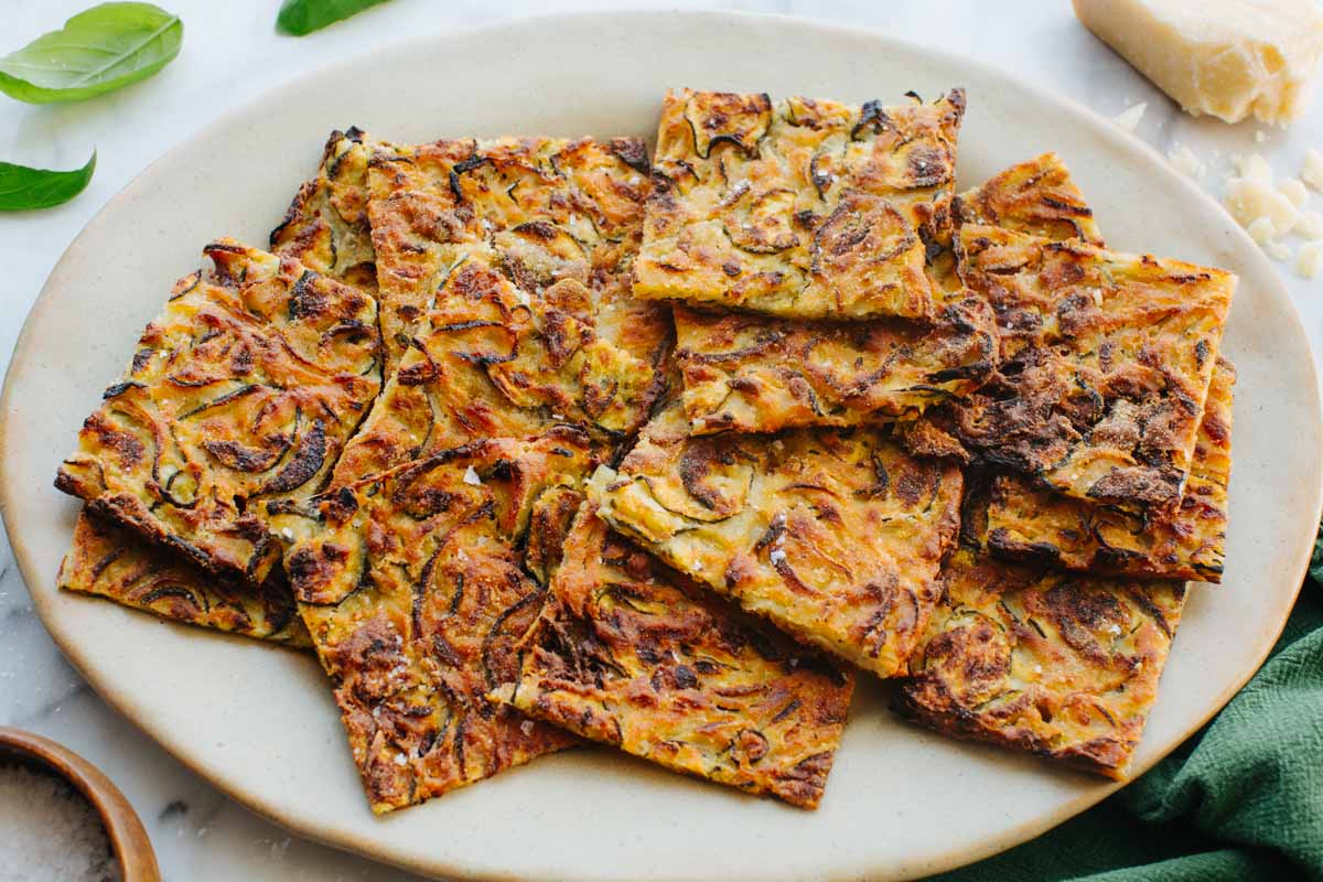 Angled view of crispy zucchini and onion scarpaccia pieces stacked on a ceramic plate, showing the thin, lacy, deeply caramelized edges.