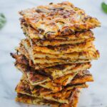 A tall stack of scarpaccia squares showing the thin, cracker-like layers, with basil leaves and grated parmesan scattered around.