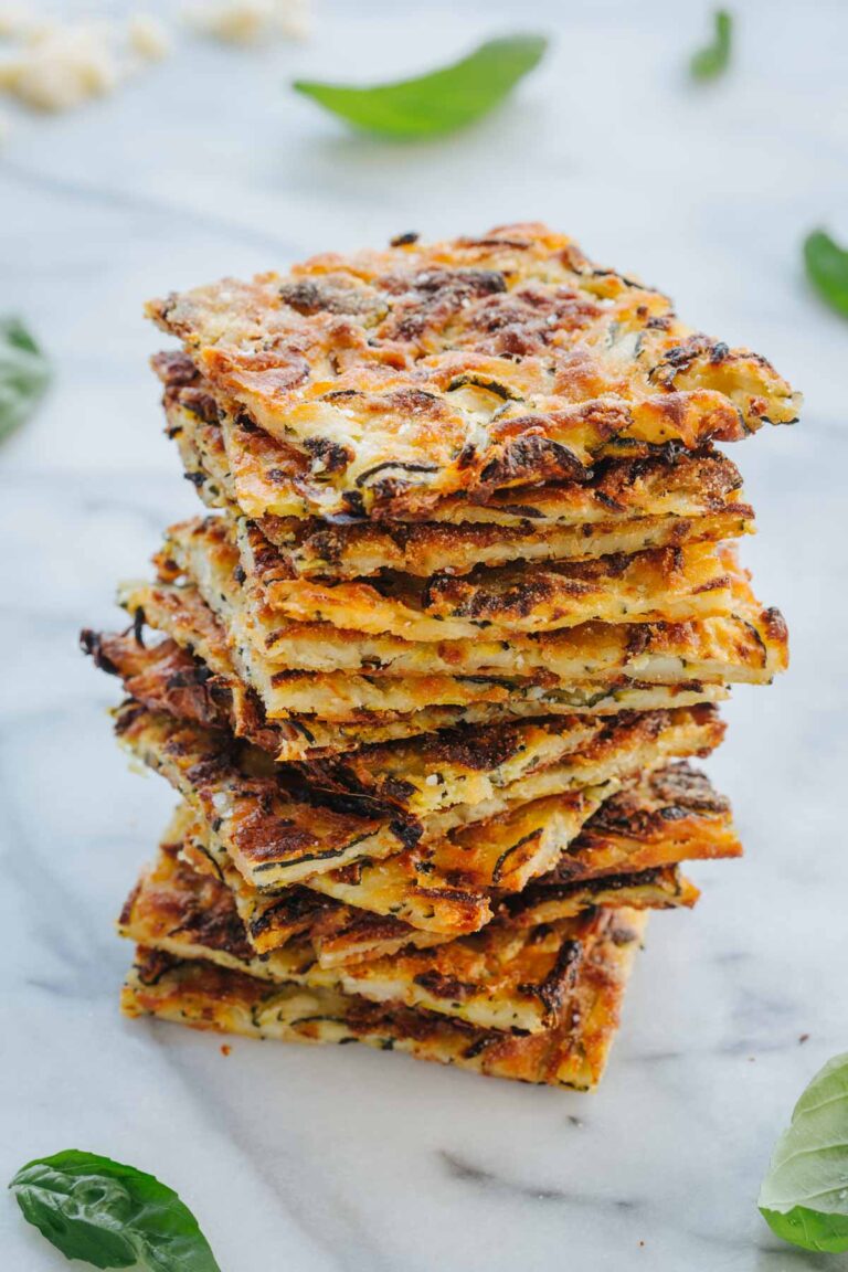 A tall stack of scarpaccia squares showing the thin, cracker-like layers, with basil leaves and grated parmesan scattered around.