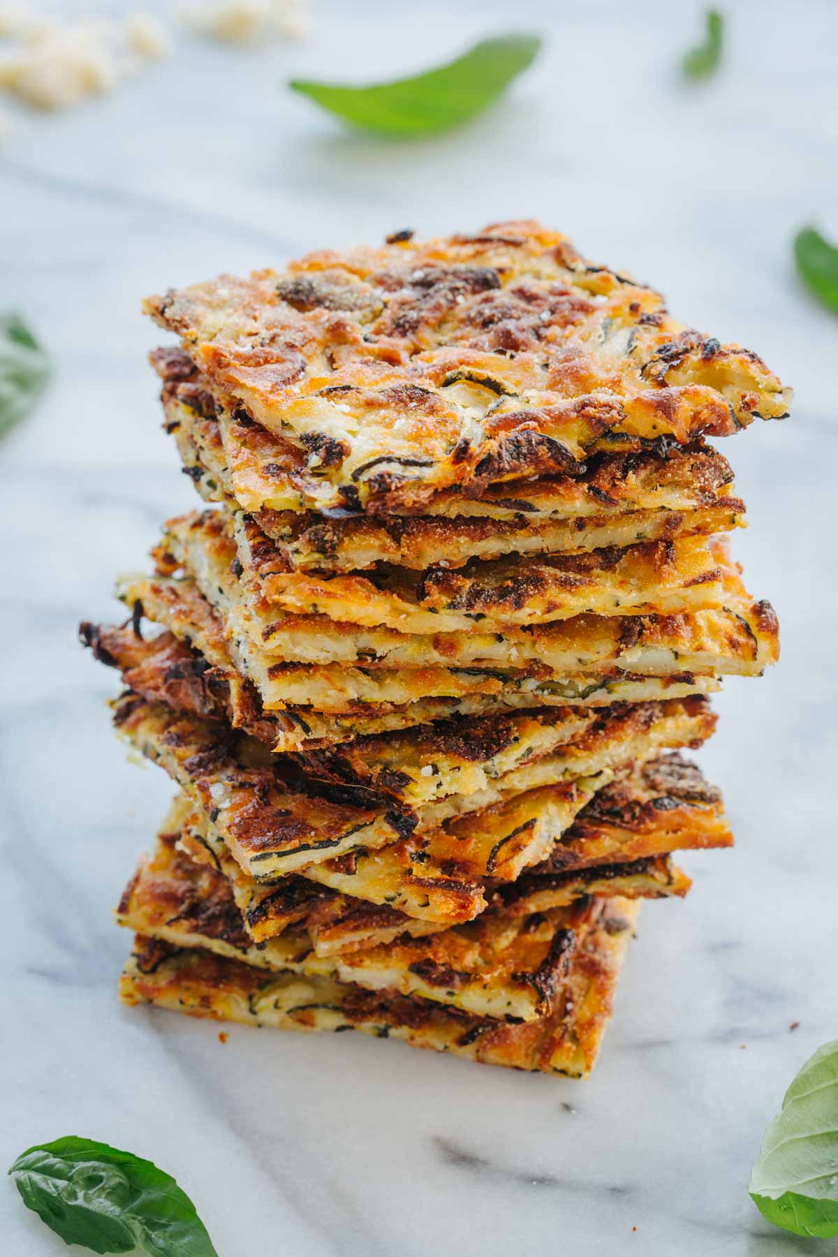 A tall stack of scarpaccia squares showing the thin, cracker-like layers, with basil leaves and grated parmesan scattered around.