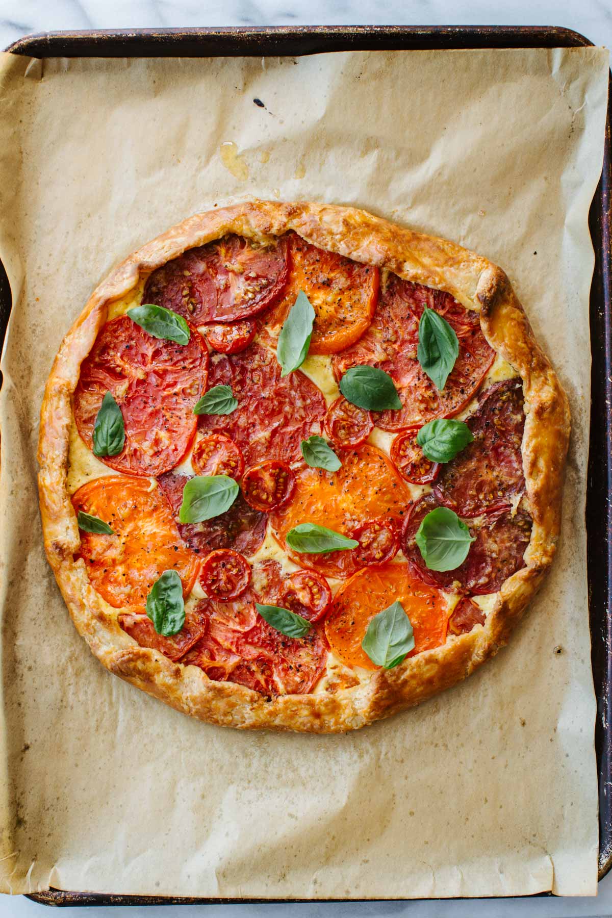 Overhead view of a whole baked tomato crostata with golden folded crust, topped with layered heirloom tomato slices in red and orange, scattered fresh basil leaves, and cracked black pepper, on a parchment-lined baking sheet.