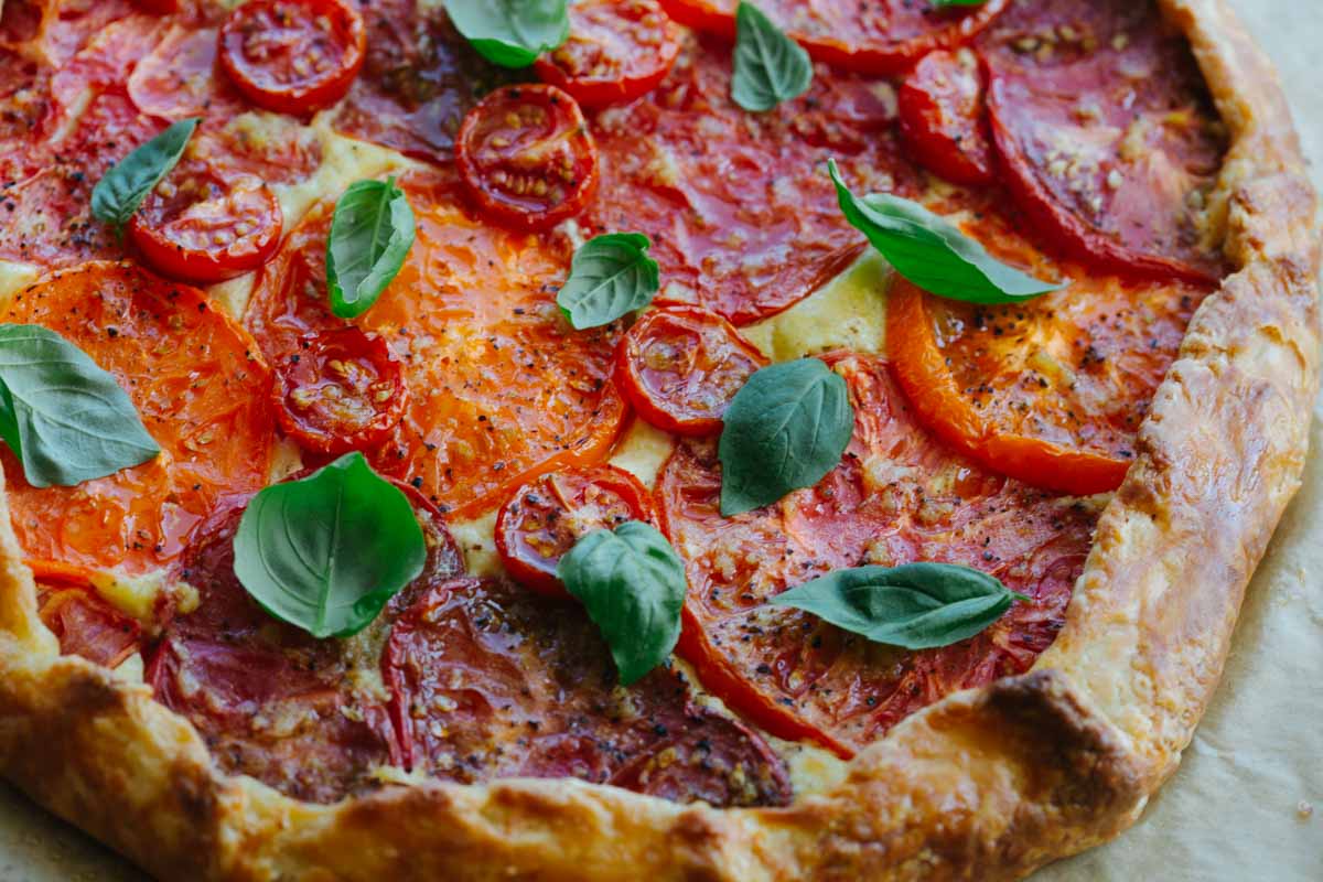 Close-up of the baked tomato crostata showing the golden, flaky crust edge and overlapping slices of red and orange heirloom tomatoes with fresh basil leaves.