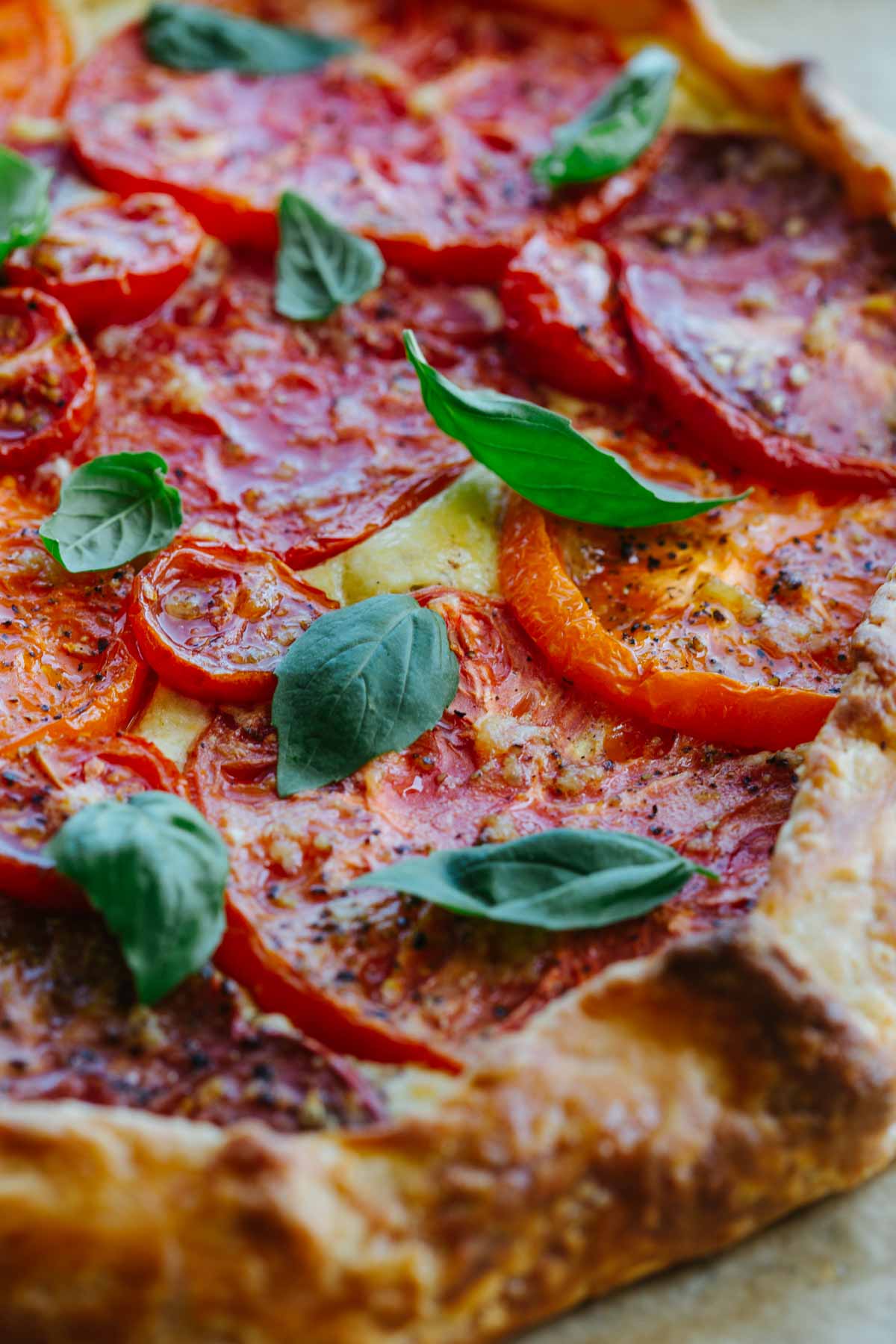 Close-up detail of the tomato crostata filling, showing glossy roasted tomato slices with cracked pepper and basil leaves against the golden pastry edge.