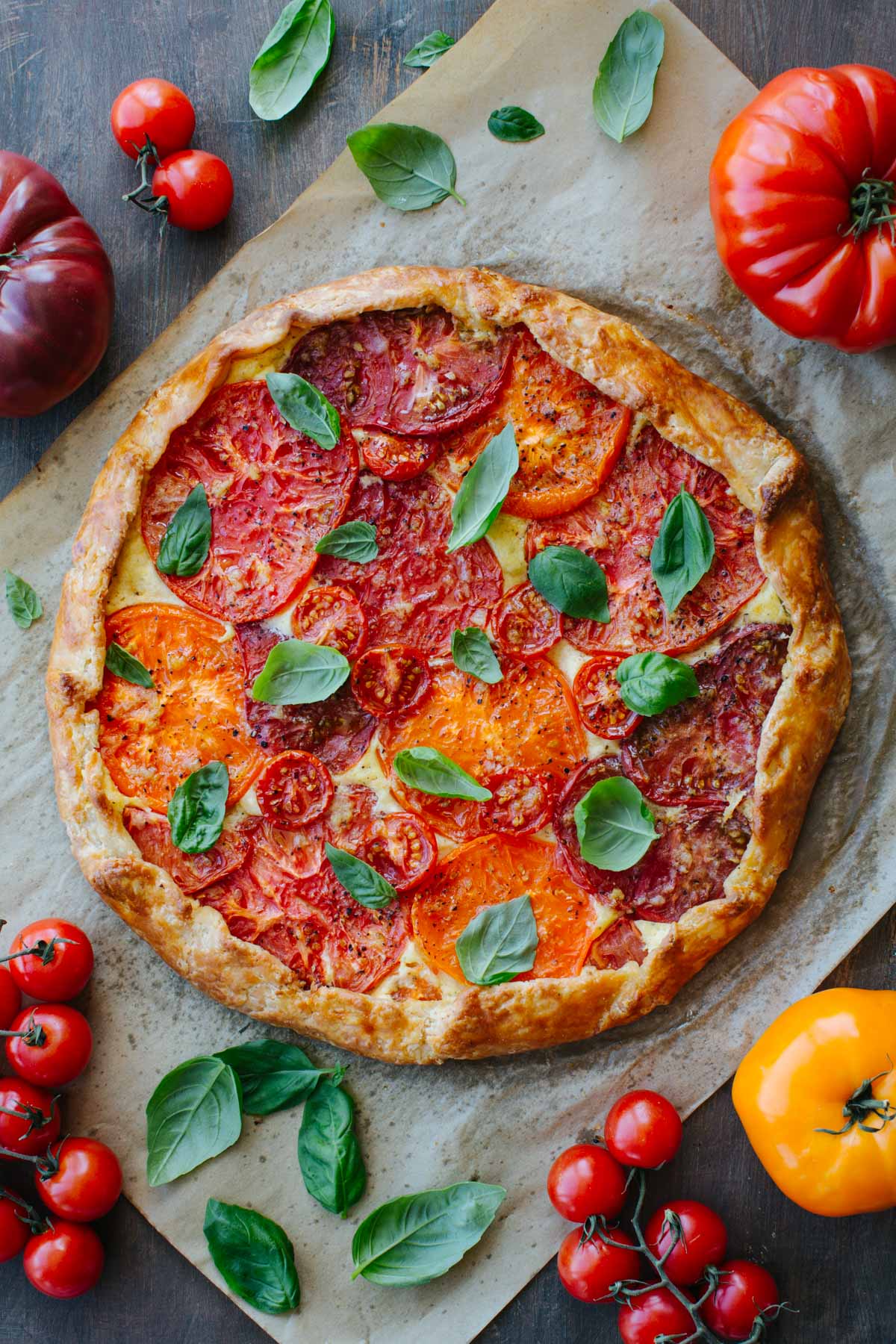 Overhead shot of the whole tomato crostata on parchment paper, surrounded by fresh whole heirloom tomatoes, cherry tomatoes, and basil leaves.
