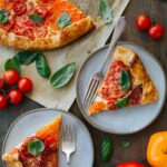 Two plated slices of tomato crostata with forks, set beside the remaining crostata, fresh basil leaves, cherry tomatoes, and a serving knife on a wood surface.