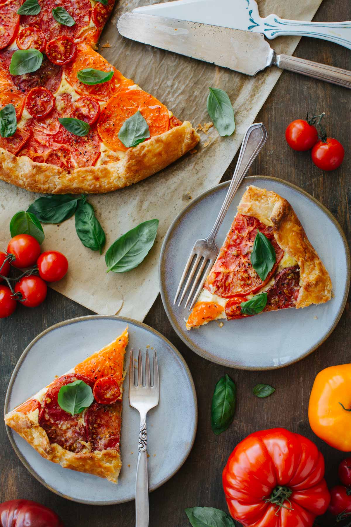 Two plated slices of tomato crostata with forks, set beside the remaining crostata, fresh basil leaves, cherry tomatoes, and a serving knife on a wood surface.
