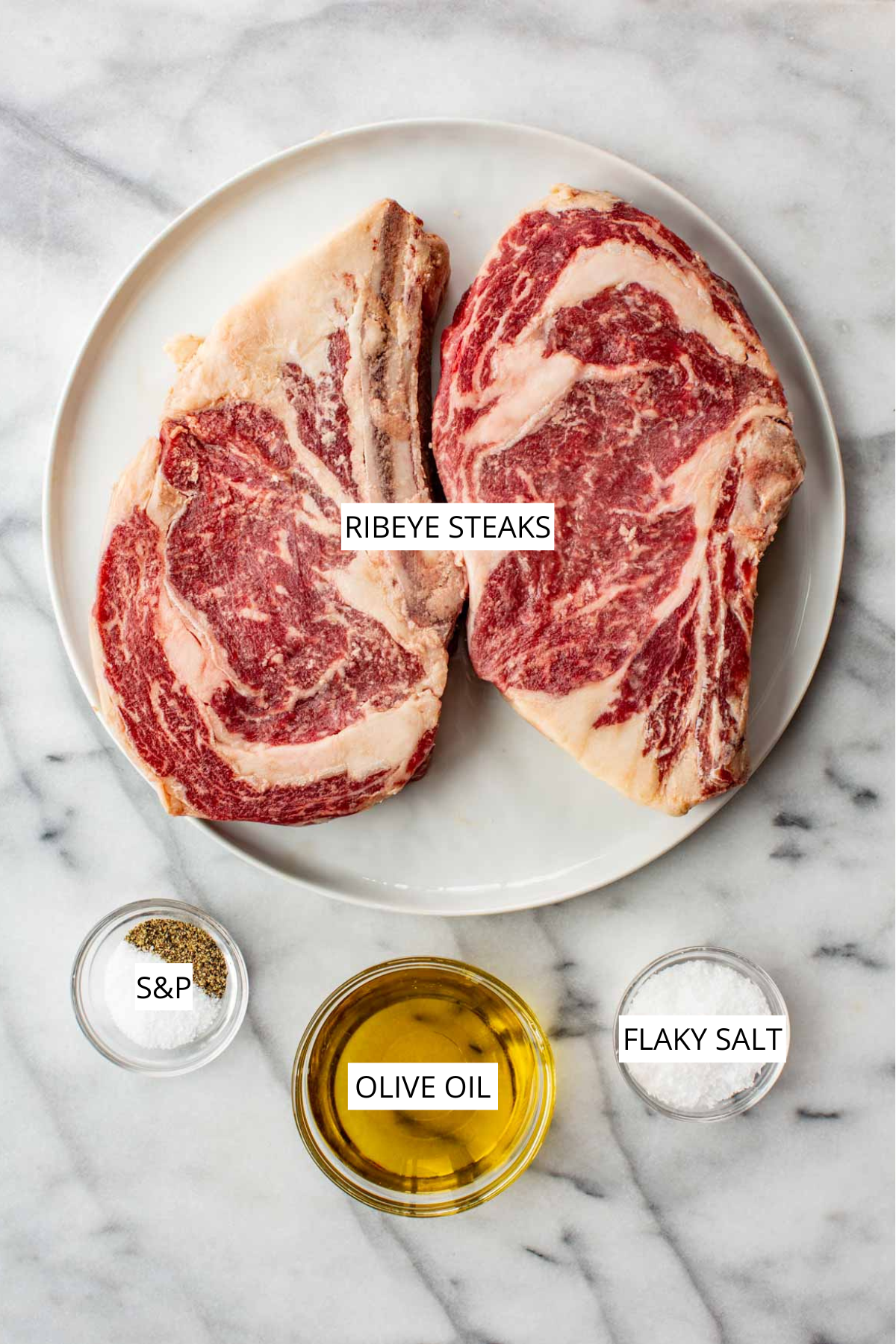 Ribeye steak recipe ingredients labeled on a marble surface: ribeye steaks, salt and pepper, olive oil, and flaky salt.