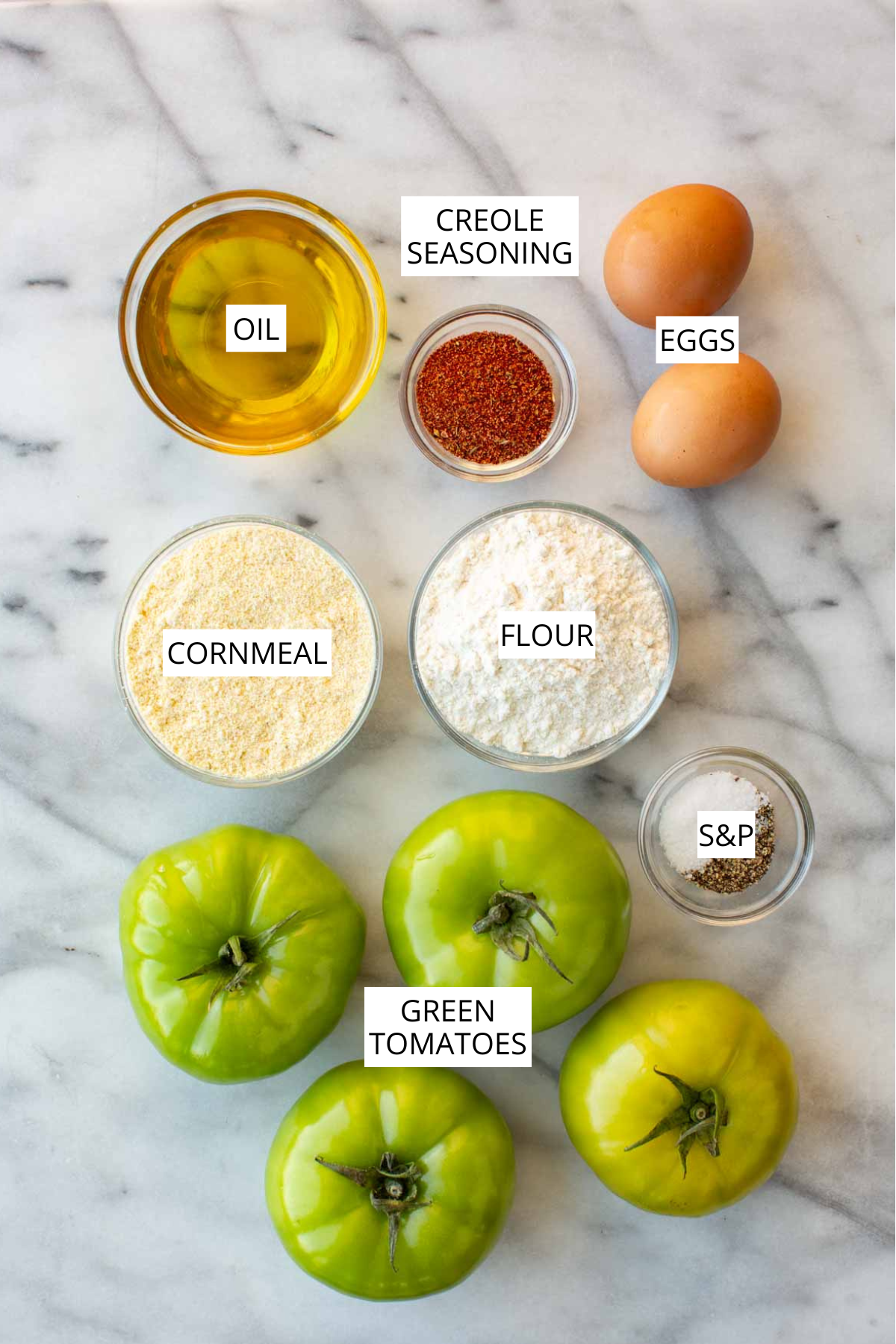Labeled overhead shot of fried green tomato ingredients: oil, Creole seasoning, eggs, cornmeal, flour, salt and pepper, and whole green tomatoes.