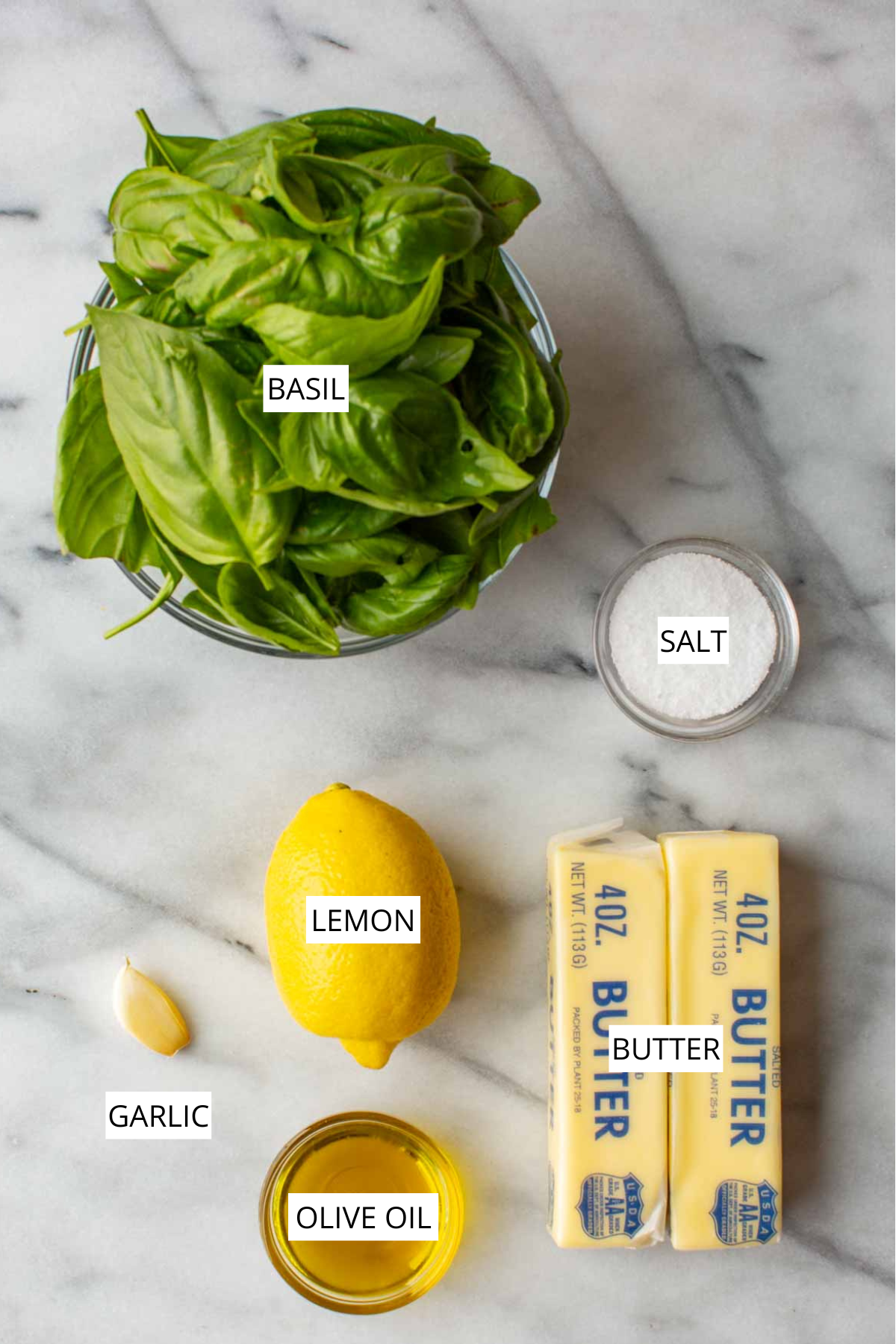Overhead shot of labeled basil butter ingredients on a marble counter: fresh basil, salt, a lemon, a garlic clove, olive oil, and two sticks of butter.