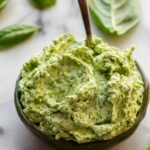 Close-up of whipped basil butter in a dark green ceramic bowl with a spoon, surrounded by fresh basil leaves.