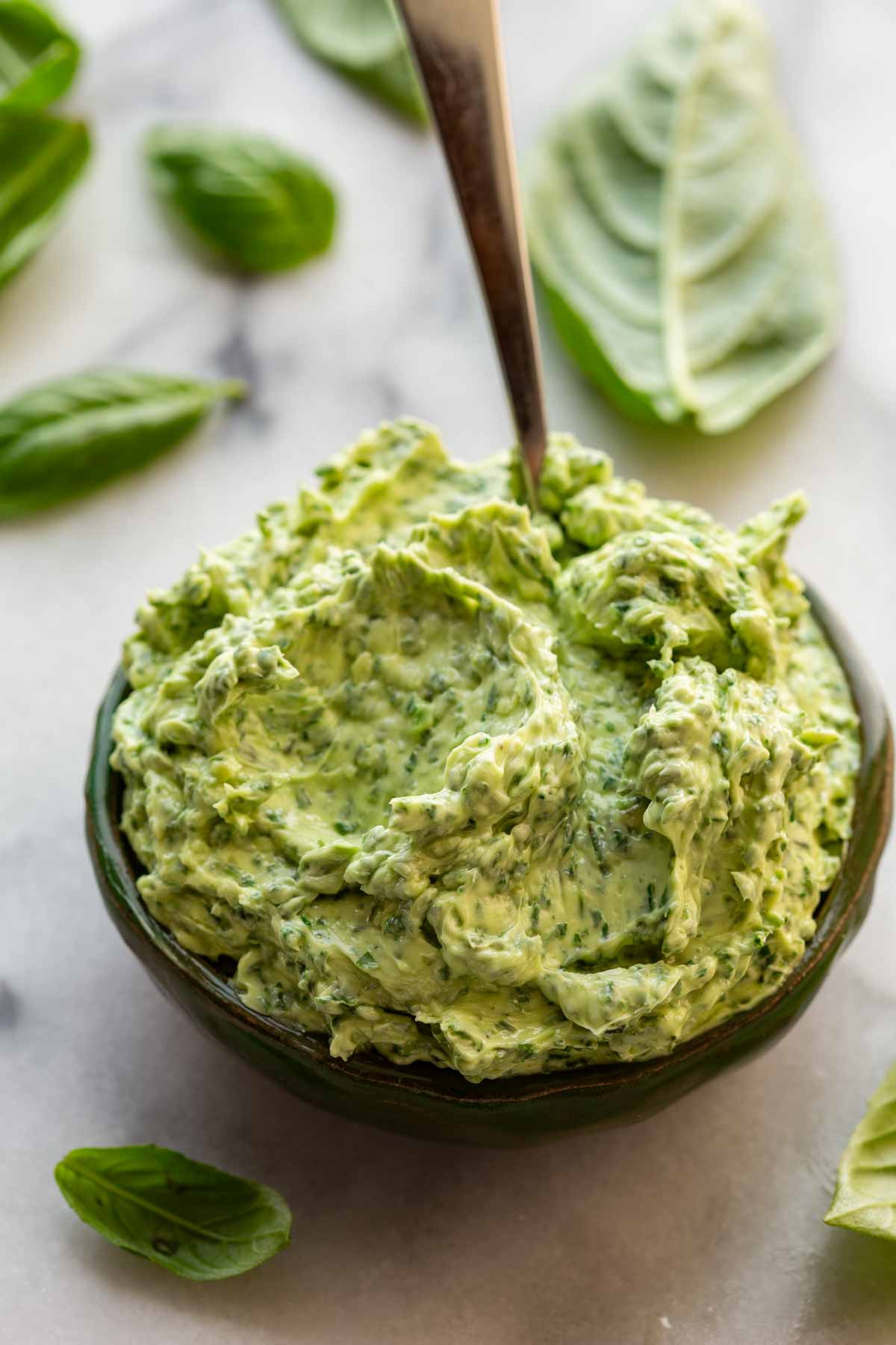 Close-up of whipped basil butter in a dark green ceramic bowl with a spoon, surrounded by fresh basil leaves.
